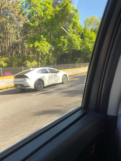 Captured through a car window, a white car is seen traveling on a road alongside lush green trees. The road is marked with a yellow median strip. The viewpoint is from within a car, framing the scene with the car's interior. The lighting suggests a bright, sunny day.