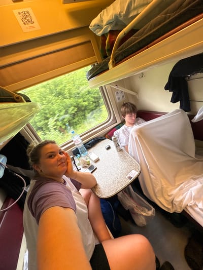 The image depicts the interior of a train compartment, with the window revealing a blur of green trees indicating movement. In the foreground, a woman with blonde hair pulled back is looking towards the camera, resting her chin on her hand. She is wearing a purple and white t-shirt. To her right, a table is visible with a water bottle, a small fan, a smartphone, and a power bank. In the background, a young boy with short brown hair is seated, wearing a white t-shirt and appearing to be looking down. He is partially obscured by a white sheet. The compartment features bunk beds with bedding, and clothing is hung on a rack. A QR code is affixed to the wall above the window. The overall lighting suggests it is daytime. The location is identified as the city of Баграш-Бигра, Russia.