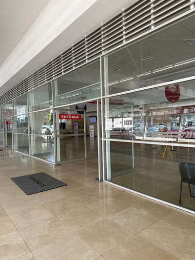 A modern building's entrance features glass walls and a door labeled "Entrada" (Entrance). A gray mat with "Bienvenidos" (Welcome) lies on the tiled floor. Reflections reveal a bright interior and ceiling details. The view through the glass includes furniture and other people inside. The architectural style emphasizes transparency and accessibility.