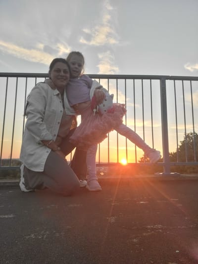 A woman and a young girl are posing for a photo in front of a metal fence with the sun setting behind them. The woman is wearing a light-colored jacket and grey pants, and the girl is wearing a purple top and a pink tutu. The sunset is casting a warm orange glow over the scene, and the sky is filled with soft, scattered clouds. The ground appears to be asphalt, and there are some scattered white debris or markings. The location is on a rooftop or elevated platform in Gillingham, United Kingdom, as indicated by the fence and the city lights visible in the distance during sunset.