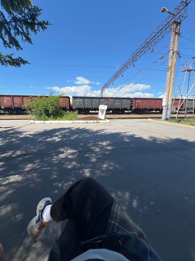 In Pereyaslavka, Russia, a person sits with their legs crossed in the foreground, their right leg extended and their left leg bent. The person is wearing black pants and white socks with athletic shoes. They are seated on a paved surface that is partially shaded by trees, casting dappled shadows. In the background, a train with several open-top cars is stopped on the tracks. The train cars are primarily dark red and grey. Overhead, power lines stretch across the blue sky, supported by a tall, industrial pole with a ladder attached. A green bush is growing between the train tracks and the paved area. The scene appears to be an outdoor train station or industrial area.