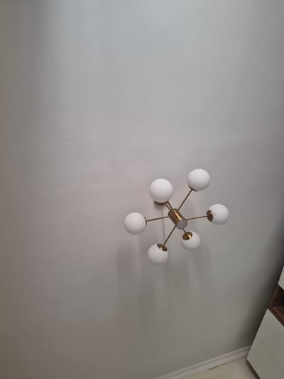 The image shows a close-up, upward view of a modern sputnik-style chandelier mounted on a plain, light-colored ceiling. The chandelier has a gold-toned metal frame with multiple arms extending outwards, each tipped with a white spherical glass globe. The globes are illuminated. To the right of the frame, a sliver of a white dresser with wooden accents is visible near the bottom edge. The lighting appears to be soft, creating subtle shadows on the ceiling. The scene is indoors, in what appears to be a residential or office space. There is no specific indication of the city of Bereke, Kazakhstan.
