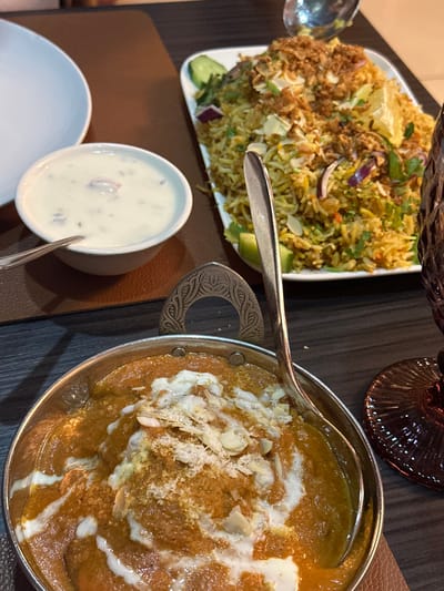 The image shows a table setting in a restaurant located at latitude 50.848 and longitude 4.374 in the city of Saint-Josse-ten-Noode, Belgium. The table is made of dark wood and has a brown placemat. There are four dishes on the table: a white bowl with a creamy white sauce, a white plate with a large portion of yellow rice with almonds and other garnishes, a silver bowl with a thick, brown curry topped with white sauce and chopped almonds, and a glass of red wine. There is a silver spoon on the table. The image captures a still life of a meal being served in a restaurant.