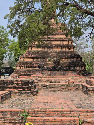 The image captures a brick structure in the Historic City of Ayutthaya, Thailand, partially obscured by trees. The brickwork appears aged and weathered, with some sections showing signs of decay and plant growth. The composition emphasizes the verticality of the structure. In the foreground, a yellow rope lies on a brick surface.