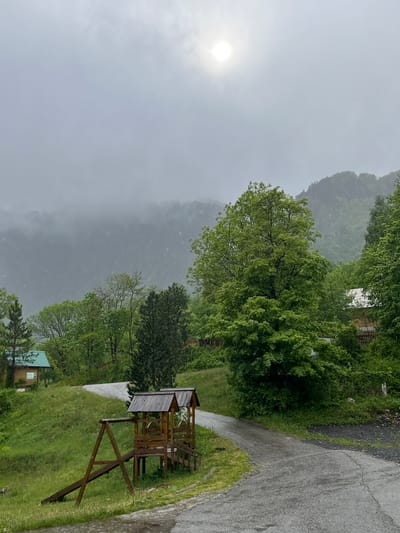 The scene is an exterior shot in Crna Poda, Montenegro. The weather is overcast, likely raining, obscuring the distant mountains with fog. A sun obscured by clouds is visible in the sky. The landscape includes green trees, a grassy slope, and a paved road. A wooden observation tower stands on the slope. A building with a green roof is situated in the lower section of the frame.