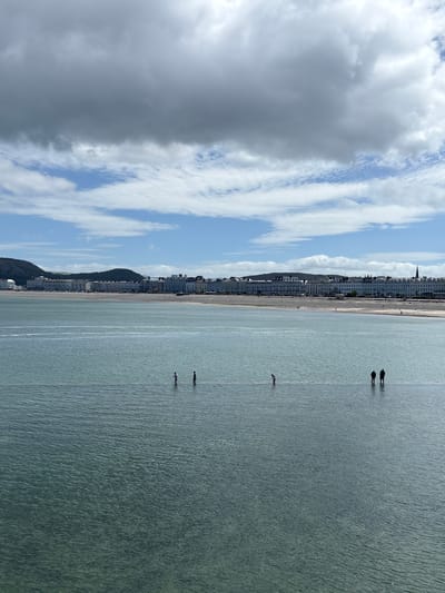 The image depicts the beach and seafront of Llandudno, United Kingdom, under a sky of mixed blue and cloudy grey. A row of buildings lines the shore. Several figures stand in the sea at various distances from the beach. The sea appears relatively calm, with slight ripples on its surface.
