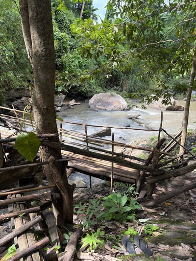 This image depicts a rustic bamboo bridge crossing a shallow, rocky river in the city of Ban Huai Takhrai, Thailand. The bridge, constructed from natural bamboo and logs, appears weathered and somewhat precarious, with a few railings missing or broken. The river flows briskly over smooth, rounded rocks, with larger boulders visible in the water and along the banks. Lush green foliage, including large-leafed plants and ferns, dominates the surrounding environment, with the dense canopy of trees casting shadows on the scene. In the foreground, a pair of dark-colored flip-flops rests on a moss-covered rock. The overall impression is one of a natural, somewhat wild setting, accessible by a rudimentary man-made structure.