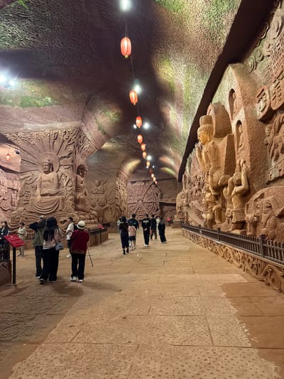 A vertically-oriented photograph shows the Dazu Rock Carvings, a series of Chinese religious sculptures and carvings, located in Dazu District, Chongqing, China.

The scene depicts a narrow corridor carved into the rock, illuminated by a string of red lanterns suspended from the ceiling. The walls are adorned with intricate carvings of Buddhist figures and scenes. People are walking along a path in the corridor, seemingly viewing the carvings.

The left wall features detailed carvings, illuminated by spotlights, while the right wall appears to be a plain stone. Above the path are more carvings and a metal railing. The floor is tiled with large stone slabs.
