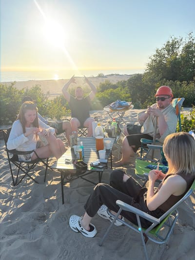 Five people are gathered on a sandy beach, likely for a picnic or small social gathering. They are seated in camping chairs around a low-lying table laden with drinks and snacks. A man in an orange hard hat and sunglasses is in the foreground, holding a cigarette. To his right, a woman with blonde hair is seated, also holding a cigarette. Two other individuals are visible in the background, one with their arms raised in a "shaka" gesture. The sun is low in the sky, casting a warm glow and long shadows across the sand, suggesting it is either sunrise or sunset. The presence of trees and a body of water in the distance indicates a natural, outdoor setting. The mood appears relaxed and casual.