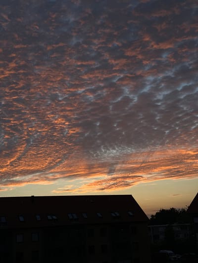 The image captures a twilight scene over Roskilde, Denmark, with the sky dominated by striated clouds illuminated by the setting sun. The lower portion of the image reveals the dark silhouettes of buildings, including a prominent red-tiled roofline with several dormer windows, suggesting an urban or suburban setting. The clouds display a mix of deep grey and vibrant orange hues, indicating the transition from day to night. The overall impression is one of a calm evening, with the architecture providing a grounded contrast to the dynamic sky. No people or specific actions are discernible.