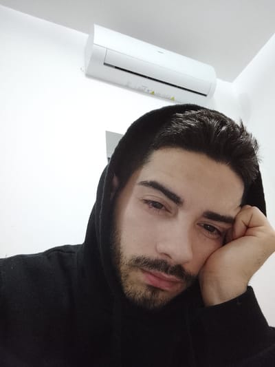A person with dark hair and a beard, wearing a black hooded sweatshirt, is resting their head on their hand. The person appears to be indoors, with a white wall behind them. An air conditioning unit is mounted on the wall in the upper part of the frame. The location is a room within the city of Fgura, Malta.