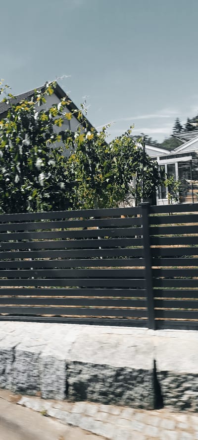The image shows a dark, horizontal slat fence in the foreground, with a stone wall or embankment below it. Beyond the fence, lush green foliage obscures part of a building with a sloped roof, and a greenhouse-like structure with a glass roof. The sky above is a hazy light blue. The scene appears to be a residential area, possibly in Winterbach, Germany, given the architectural style of the buildings and the presence of what looks like a vineyard or garden. The overall impression is one of a well-maintained outdoor space with a naturalistic element.