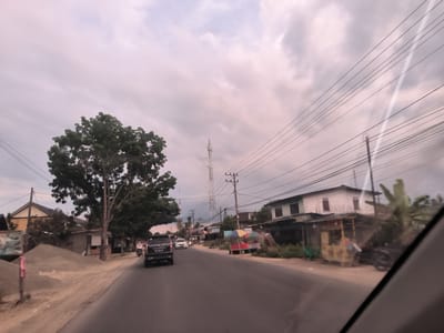 The image depicts a street scene in Keudebirem, Indonesia. A two-lane paved road stretches into the distance, with a black SUV traveling on the left lane. A large, full tree stands to the left of the road. On the right, there are buildings, including a two-story white structure with a lower attached structure. Utility poles with numerous wires span across the street overhead. Further down the road, a cell tower rises into the overcast sky. There are parked vehicles and people visible along the roadside in the distance. A sandy area with a pile of materials is located on the left side of the road.