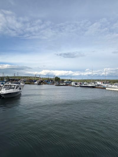 The image shows a marina in Molodezhnyi, Russia, under a cloudy sky. Several boats of various sizes are docked along a pier. In the foreground, a white motorboat with a blue stripe is prominent. In the background, there are more boats, some with masts, along with buildings and structures that appear to be part of the marina's infrastructure. Green fields and trees are visible on the horizon. The water is calm with ripples. No people are visible in the image.
