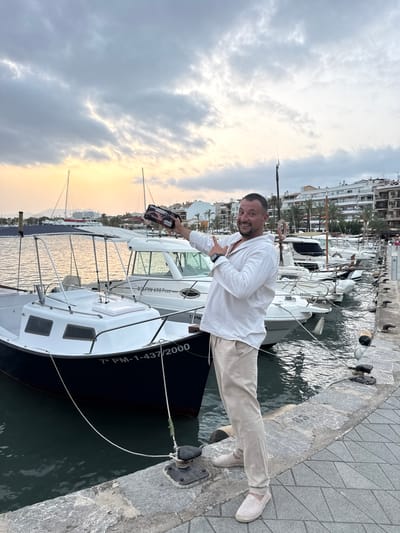 A man in a white linen shirt and beige pants stands on a stone pier in Port D'alcúdia, Spain, holding a small, dark object with a patterned design in his right hand. He is gesturing with his left hand towards the object. Behind him, several white boats are moored in the water, with their masts and rigging visible. In the background, buildings line the waterfront, and palm trees are scattered among them. The sky is cloudy, with a soft glow of sunset on the horizon. The water is calm, reflecting the sky and the boats. The scene appears to be a harbor at dusk.
