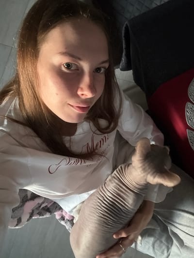 A young woman with brown hair pulled back is holding a hairless cat in her lap. She is wearing a white sweatshirt with a maroon graphic and gray sweatpants. The cat is light brown and has visible wrinkles. The woman is looking at the camera with a slight smile. In the background, a dark blanket and a red object with a decorative pattern are visible. The scene appears to be indoors, possibly in a residential setting. No specific location in Novaya Ivanovka, Russia, can be determined from the image.