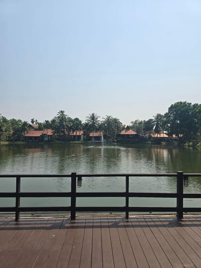 The image depicts an outdoor scenic view across a body of water, located in Ban Phaya Kot Ana Kaeo, Thailand. In the foreground, a dark brown wooden deck with parallel planks is visible, featuring a black wooden railing that spans the lower third of the frame. An industrial-style wall sconce with a cage cover is affixed to a vertical post of the railing on the right. The midground features a calm, dark green body of water, exhibiting minor surface ripples and a multi-jet water fountain actively spraying near the left-center. The background reveals the far bank lined with several buildings characterized by reddish-brown tiled roofs and lighter-colored walls, largely integrated within a dense landscape of green tropical foliage, predominantly palm trees. A small bridge extends across a section of water or land on the right side of the far bank. The upper portion of the image is dominated by a clear, pale blue sky. No human or animal subjects are present. The scene is brightly illuminated by natural light, casting distinct shadows from the railing onto the deck.