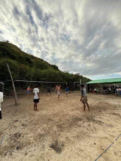 The image shows a beach volleyball game in Rinca Village, Indonesia. A volleyball net is visible with several people standing on the sand playing or preparing to play. A forested hill rises in the background to the left. Behind the volleyball court, further in the background, is a covered area with people seated and watching. The sky is cloudy. The beach sand is light-colored.