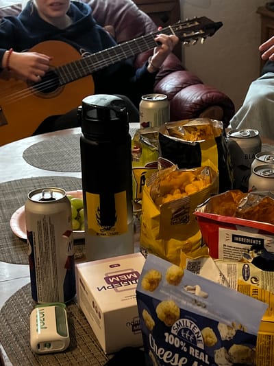 A gathering with food and drink appears on a table. A person is playing a guitar. The food includes potato chips in several bags. Drinks include several cans of beer or similar beverages. There's a box of pills. A small white electronic device sits on the table. The location is implied through a "Doamn Tours" label to be in or around Kalnciems, Latvia.