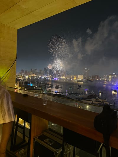 A nighttime view showcases fireworks exploding above a cityscape and waterway. Tall buildings are illuminated along the water's edge, where boats are visible. The foreground features a partial view from an elevated position, including a wooden railing, a black backpack, and a glimpse of someone's white clothing. The overall ambiance is festive.