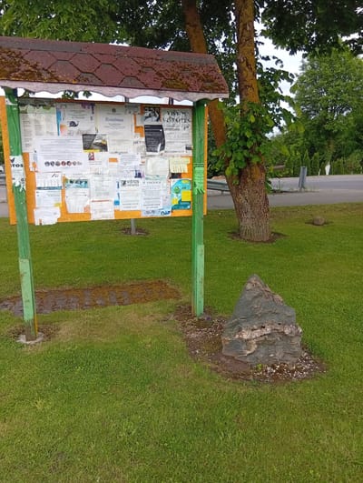 The image depicts an outdoor bulletin board in Micaičiai, Lithuania, displaying various notices and advertisements. The bulletin board is made of wood with a red, moss-covered roof and is supported by two green wooden posts. It is situated on a grassy area, with a large, uniquely shaped rock positioned in front of it. Behind the bulletin board, a large tree with thick branches and green leaves dominates the background. The area surrounding the bulletin board is covered in lush green grass, and in the distance, a road with a metal guardrail is visible, along with more trees and foliage. The scene appears to be a public space, likely within the village of Micaičiai.