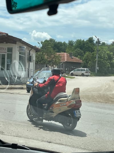 A photo taken from inside a car shows a man on a Suzuki motorcycle with license plate BB 5114 in Tsar Kaloyan, Bulgaria. The man wears a red jacket and helmet. The motorcycle is parked on the side of the road in front of a building with the word "TEKA" visible, and a white van is parked further down the street. The environment appears to be a residential area.
