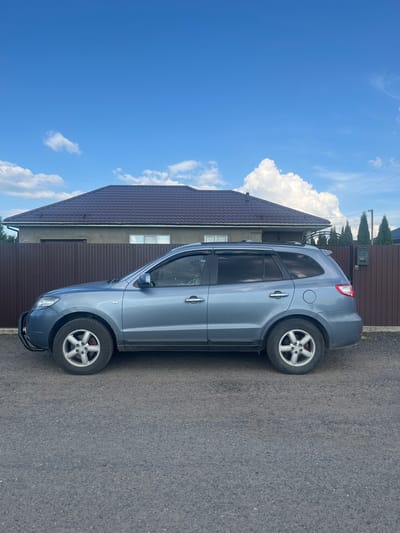 This media file depicts a gray SUV parked on an asphalt surface next to a building. The SUV is positioned facing the viewer's right. The building has a dark brown, tiled roof and brown siding. In the background, a brown wooden fence can be seen, with some trees and a mailbox. The sky is bright blue with scattered white clouds, suggesting it is daytime and the weather is fair. The scene appears to be in a rural or suburban setting. There are no people visible in the image. The image is oriented vertically. No visible text is present.