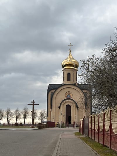 The image depicts an Eastern Orthodox church in Абухаўскі сельскі Савет, Belarus, on a cloudy day. The church features a prominent golden dome and a cross on top. The building is adjacent to a paved road. A wooden fence with a distinctive pattern runs alongside the road, parallel to the church. Further down the road, multiple large crosses stand in a row. Leafless trees are visible in the background.