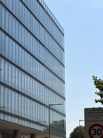 This media file captures a low-angle view of a modern building with a sloped, glass-paneled facade. The building appears to be contemporary, with its sleek design and reflective surfaces. The sky above is a clear, pale blue, suggesting a bright day, possibly late morning or early afternoon. A few wispy clouds are visible, and a faint contrail from an airplane stretches across the sky.

At the top right of the frame, a street sign is partially visible. It is mounted on a pole and displays text in French, along with a speed limit sign indicating "30". The text on the sign appears to be a regulation, with phrases like "LE CODE DE LA ROUTE S'APPLIQUE" (The Highway Code applies) and "LA VITESSE EST LIM" (The speed is lim[ited]). Above and to the left of the sign, the green leaves of a tree are visible, providing a natural contrast to the urban architecture.

The overall scene is one of an urban environment, likely a business district or a public space, given the presence of the speed limit sign and the architectural style of the building. The image is static, focusing on the architectural elements and the sky. There are no people visible in the frame. The lighting suggests it is daytime, with the sun casting reflections on the glass. The location context of Rungis, France, indicates a specific geographical setting for this observation.