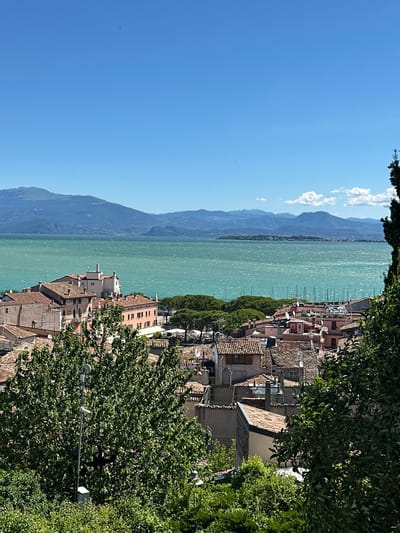 The image shows a panoramic view of the town of Desenzano del Garda, Italy, on a clear, sunny day. In the foreground, dense green foliage obscures parts of the view, with the rooftops of buildings peeking through. The buildings are characterized by terracotta tile roofs and pastel-colored walls, typical of Mediterranean architecture.

In the mid-ground, the town extends towards a large body of water, which appears to be Lake Garda, identified by the surrounding mountains. The water is a vibrant turquoise color, with gentle ripples on its surface. Numerous sailboats are docked or sailing on the lake, their masts creating a delicate white pattern against the water.

The background is dominated by a range of majestic mountains, their peaks softened by distance and a light haze. The sky is a brilliant, cloudless blue, with a few wispy white clouds visible in the distance. A small dark object, possibly a bird or a distant drone, is visible in the upper left portion of the sky. The overall impression is one of a serene and picturesque lakeside town on a beautiful summer day.