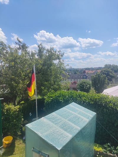 This is an outdoor, eye-level, full-shot photograph taken on a sunny day in Chemnitz, Germany. The scene is a backyard with lush greenery and a view of residential buildings in the background. In the foreground, a turquoise greenhouse with a translucent cover is positioned. To the left of the greenhouse, a yellow bucket sits on the grass. A German flag, with its black, red, and yellow stripes, waves proudly from a flagpole in front of a large, leafy tree. The sky above is a vibrant blue, dotted with fluffy white clouds, indicating fair weather. The sunlight suggests it is likely daytime, perhaps late morning or afternoon. The overall mood is peaceful and suggests a typical summer day in a suburban or urban garden setting. There is no visible text in the image.