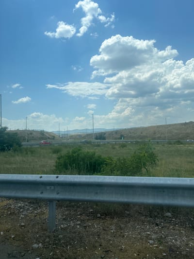 The image depicts a roadside scene under a bright, partly cloudy sky. A silver metal guardrail runs across the foreground, with its supporting posts visible. Beyond the guardrail, a dry, grassy field stretches towards a highway where two cars are visible: a red car in the left lane and a white car in the right lane. The highway is bordered by a grass embankment dotted with sparse trees and tall, thin lampposts. In the distance, rolling hills with dry vegetation are visible, and the horizon is hazy. The environment appears to be arid, with dry grasses and sparse greenery. The location is identified as Barakovo, Bulgaria. There are no people clearly visible in the image.