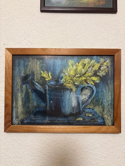 A framed pastel drawing of a blue watering can with yellow foliage is displayed on a textured white wall. The watering can is on a dark blue surface, possibly a table or shelf. The background is a mix of dark blue, green, and yellow, suggesting a textured or abstract environment. The drawing is in landscape orientation and has a thin wooden frame. Above and to the right of the main artwork, a portion of another framed picture with a dark frame is visible against the white wall. The image is taken from a slightly elevated angle, looking down at the artwork. There are no people or animals present in the image. The overall scene is still and appears to be a domestic interior.