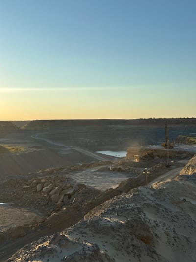 This media file captures a wide, panoramic view of an open-pit mine, likely a quarry, in Tatsinskaya, Russia. The scene is dominated by the layered rock formations and excavated earth, indicative of large-scale mining operations. In the mid-ground, a yellow mining vehicle, possibly a drill rig or loader, is visible. A body of water, likely collected rainwater or a pond, is situated at the bottom of the quarry. The overall impression is one of industrial activity against a natural, rugged landscape.
The time of day appears to be late afternoon or early evening, given the warm, golden light illuminating the upper sections of the quarry and casting long shadows. The sky transitions from a clear, pale blue on the left to a hazy, yellowish hue towards the horizon on the right, suggesting a sunset or sunrise. The weather appears clear, with no visible precipitation or overcast conditions.
Notable details include the visible stratification of the rock walls, suggesting different geological layers that have been extracted. Winding roads or tracks are visible throughout the quarry, indicating the movement of heavy machinery. The texture of the exposed rock surfaces is rough and varied, with some areas showing sharp, angular fragments and others appearing more weathered and eroded. The image conveys a sense of scale and the immense effort involved in extracting resources from the earth. No visible text is present in the media file.