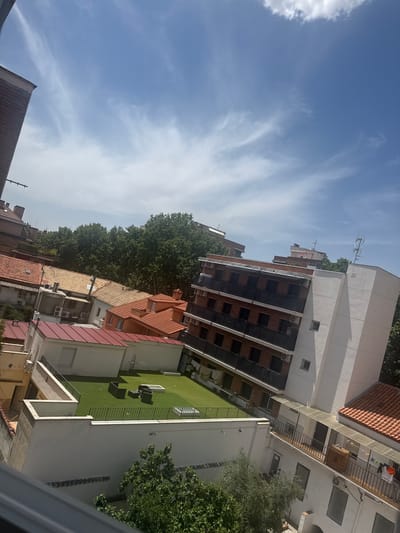 This image captures an aerial view of a residential area in Alcalá de Henares, Spain, under a partly cloudy sky. The scene is dominated by buildings with tiled roofs and modern apartment complexes. In the foreground, a rooftop area features artificial grass, a small seating area with dark furniture, and a low white wall with a black railing. Adjacent to this is a white building with a red metal roof, several windows, and an air conditioning unit. Further back, more buildings with orange tiled roofs and white walls are visible, interspersed with trees. A larger, modern building with a white facade and multiple balconies with dark railings occupies the right side of the frame, while a brick building with similar balconies is positioned diagonally in the center. Various air conditioning units are visible on the exteriors of the buildings. Trees are scattered throughout the scene, providing greenery amidst the urban landscape. The overall impression is of a sunny day in a built-up urban environment.