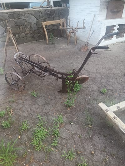 An old, rusty plow is the central focus of this outdoor scene, which appears to be in a courtyard area. The plow, with its two large metal wheels and a single furrowing blade, rests on damp, dark soil. Green weeds and small plants are growing around it, indicating a somewhat unkempt environment.
Behind the plow, a rough-hewn stone wall forms a backdrop. To the right of the wall, a white brick structure, resembling an outdoor oven or fireplace, is visible. Leaning against this brick structure are a shovel and a long-handled tool.
Other wooden agricultural implements and structures are scattered around the area, suggesting a historical or rustic display. A wooden structure with curved supports and what look like antlers or branches protrudes from the ground to the left of the plow. Further to the right, a part of a wooden frame structure is visible in the foreground.
The overall impression is that of an outdoor exhibit or storage area for old farm equipment, possibly at a historical site or museum. The lighting suggests it might be overcast or late in the day. There are no people present in the image.