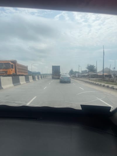 The image depicts a multi-lane highway in Akodo, Nigeria, viewed from the interior of a vehicle through a severely cracked windshield. The vehicle's dark, textured dashboard is visible at the bottom of the frame. The roadway features at least two lanes of travel, demarcated by white dashed lines, and a concrete median barrier with alternating black and white stripes.

In the left lane, an orange dump truck, heavily loaded with light-colored, granular material, is positioned in the mid-ground, appearing stationary or moving slowly. In the right lane, a dark-colored sedan is observed ahead, followed closely by a dark flatbed truck.

The roadside on the right comprises an unpaved shoulder leading to a strip of dry, light-colored soil with sparse vegetation, including several thin trees and a distinctive palm-like tree. Numerous utility poles of varying heights are present along this strip. Beyond them, several rudimentary structures, consistent with small shops or dwellings, are visible, constructed from materials such as corrugated metal. Piles of various indeterminate materials are clustered near these structures.

The sky is overcast with a continuous layer of grey and white clouds. The foreground windshield exhibits extensive fracturing, including prominent horizontal cracks across the lower central portion, with additional cracks extending diagonally and vertically, particularly towards the right side. A dark, irregular patch is affixed or painted onto the lower right corner of the windshield.