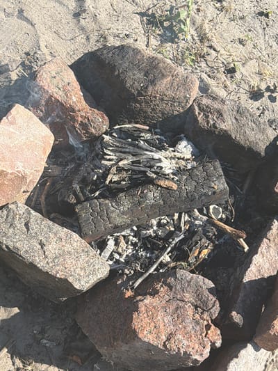 This image captures a close-up view of a smoldering campfire pit, constructed from rough-hewn stones, on a sandy surface with sparse dry vegetation and weeds. The pit contains the remnants of burnt wood, ash, and embers, with a few thin tendrils of smoke rising from the center. The surrounding stones are varied in color and texture, some appearing reddish and others dark gray and speckled. The lighting suggests it is daytime, with shadows cast by the rocks. The scene is devoid of human presence. The location is identified as the city of Lebedivka, Ukraine.