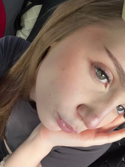 The image is a close-up, overhead selfie of a young Caucasian female. She has light brown hair and fair skin with some blush on her cheeks. Her eyes are green and she is wearing mascara. Her lips are slightly parted and appear to be glossed. Her right hand is resting on her chin, and she is wearing a gray shirt. The background is out of focus and appears to be a car interior, with a patterned seat and black upholstery. The specific location in Bairak, Russia, cannot be determined from the image.