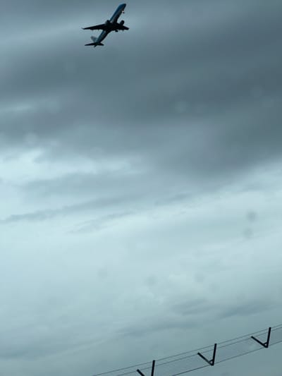 The image shows a low-angle view of an airplane in flight against a gray, overcast sky. A chain-link fence is visible in the upper right corner. It is impossible to determine the location of the image within Broadland, United Kingdom with the provided information. No people are visible.