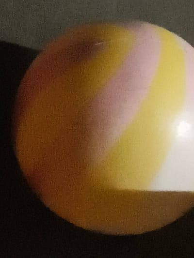 The image is a close-up shot of a ball with swirling colors of orange, yellow, and pink. The ball is spherical and appears to be smooth. The background is dark and out of focus, suggesting the ball is the main subject. There is a bright highlight on the right side of the ball, indicating a light source. The image quality is grainy. No specific location in Ubitpayaitek, Indonesia, can be determined from the provided image.