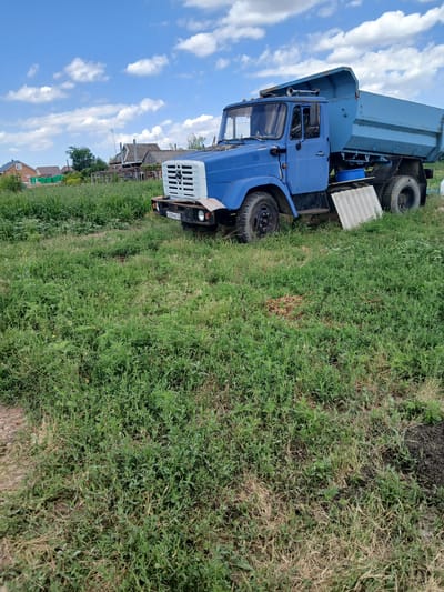 A blue dump truck is parked on the side of a grassy area, likely in a rural setting. The truck has a large, light blue dump bed that is elevated. The cabin of the truck is also blue with a white grille. The tires are black and appear to be in good condition. The scene is set outdoors, with a bright blue sky and fluffy white clouds visible in the background. The grass is green and lush, with some patches of dry, brown grass. In the distance, there are some houses and fences, suggesting a residential area. The overall impression is one of a bright, sunny day. The license plate of the truck is visible and reads "R544PO75". There are no people visible in the image. The time of day appears to be daytime, given the bright sunlight.