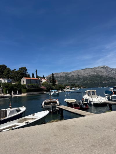 The image shows a sunny day with a clear blue sky over a harbor in Grad Korčula, Croatia. Several boats are docked in the calm water. In the foreground, a paved walkway lines the edge of the harbor, with a white boat partially resting on the shore. Further out in the water, more boats are visible, including a white motorboat with "Pinka" written on its side and another boat labeled "BOAT TOUR". The background features lush green trees and Mediterranean-style houses with red tile roofs, leading up to a rocky, tree-covered mountain. The scene depicts a typical coastal village atmosphere.