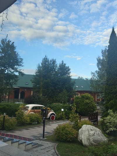 This media file captures an outdoor scene with a clear blue sky dotted with fluffy white clouds. The perspective is slightly tilted. In the foreground, a paved walkway with a geometric pattern leads through a lush garden. Several trees and shrubs with varying shades of green foliage are visible. A distinctive feature is a large, rounded rock with swirling patterns, placed prominently in the garden.
Further back, a brick building with a green tiled roof and dormer windows can be seen, suggesting a residential or possibly a commercial property. A quaint, vintage-style vehicle, possibly a decorative piece or an art installation, sits beside the walkway. It's white with orange accents and a red wheel.
The scene appears to be set during the daytime, with bright sunlight casting shadows. The overall atmosphere is serene and picturesque, evoking a sense of a well-maintained garden or park. The location is identified as Tsibino, Russia. No people are visible in the frame, and there is no discernible text within the image.