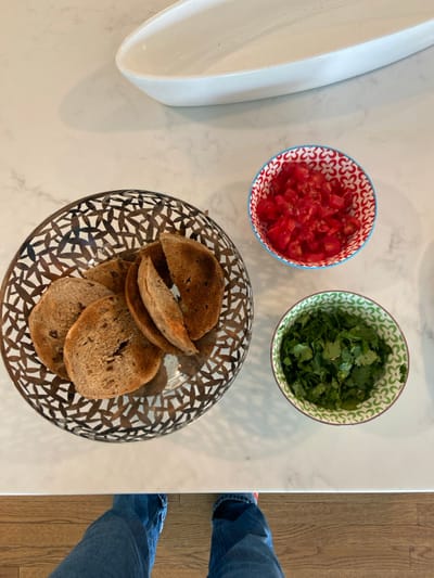 A white countertop in Rye, New York, is the setting for a scene containing three dishes: a silver dish with six pieces of toasted bread, a red and blue patterned bowl filled with diced tomatoes, and a green and white patterned bowl filled with chopped cilantro. The top of the countertop is white, with a light brown wooden surface below. Only the bottom portion of a pair of blue jeans is visible in the lower portion of the image, indicating the photographer is standing above the countertop.