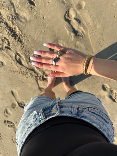 A high-angle forensic shot depicts a person standing barefoot on light-brown sand at a beach in Manhattan Beach, United States. The upper thighs and right arm are visible. The person wears light-wash distressed denim shorts over black undergarments. Their bare feet are partially visible, positioned in the lower-middle of the frame, surrounded by various footprints in the sand. The right hand, extended forward and slightly to the left, is prominently featured. It displays long, squared-off fingernails with a white ombré design. On the index finger is a silver-toned signet ring with a dark circular emblem. On the middle finger is a gold-toned signet ring with a yellow, possibly sun-like, design. The right wrist is adorned with two bracelets: a thick, ridged gold-toned bangle and a thinner gold-toned chain. Bright sunlight casts distinct shadows, particularly from the person's hand and body onto the sand to the left.