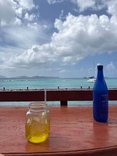 This image, captured in Spanish Town, British Virgin Islands, presents an outdoor waterfront scene under a bright, partly cloudy sky. The foreground features a reddish-brown wooden surface, likely a table or bar. On this surface, a clear glass mason jar, containing a yellow liquid with ice and a clear plastic straw, is positioned on the left. To its right, a blue glass bottle with a dark blue cap stands, exhibiting condensation and a label reading 'SARATO EXQUISITE Effervescent.' A dark reddish-brown wooden railing, adorned with a string of white fairy lights, crosses the midground horizontally. Beyond the railing, the turquoise ocean extends into the distance, with several boats, including at least two sailboats, visible. Distant landmasses are silhouetted against the horizon under a sky filled with cumulus clouds. No individuals are present in the frame.