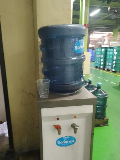 A water cooler is positioned next to a light yellow wall. The water cooler has a large blue water bottle on top, with the brand "Bonanza" visible. To the left of the water cooler is a transparent plastic cup. Below the water bottle, the cooler itself is white with two dispensing taps, one orange and one silver. A blue oval sticker with the "Bonanza" logo is also present on the cooler. In the background, there are stacks of green crates and a green floor. The scene appears to be inside a warehouse or factory, consistent with a location in Sukaresmi, Indonesia, a known industrial area. There are no people visible in the image.