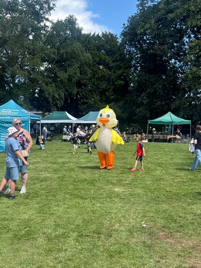 A bright, sunny day event is taking place on a grassy field. In the foreground, a person in an inflatable duck costume, with yellow body and orange legs and beak, stands centrally. To the left, a woman wearing sunglasses and a floral top stands next to a child in a blue t-shirt and shorts. Several other people, including children and adults, are scattered across the field, some interacting, others observing. In the background, there are several gazebos and tents, with activities and stalls set up. Tall, mature trees form a backdrop to the scene. The event appears to be a community gathering or fair, possibly held in a park or open space within Leominster, United Kingdom.