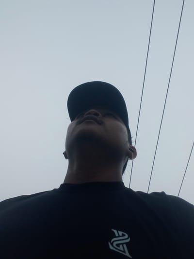 A low-angle, close-up, outdoor, medium shot shows a person wearing a black t-shirt and a black baseball cap. The person is looking upwards, and their face is partially obscured by the cap. The background is a light gray sky with thin, dark, parallel lines that appear to be power lines. The person's t-shirt has a white logo on the left chest. The scene is in Kubang Jaya, Indonesia.